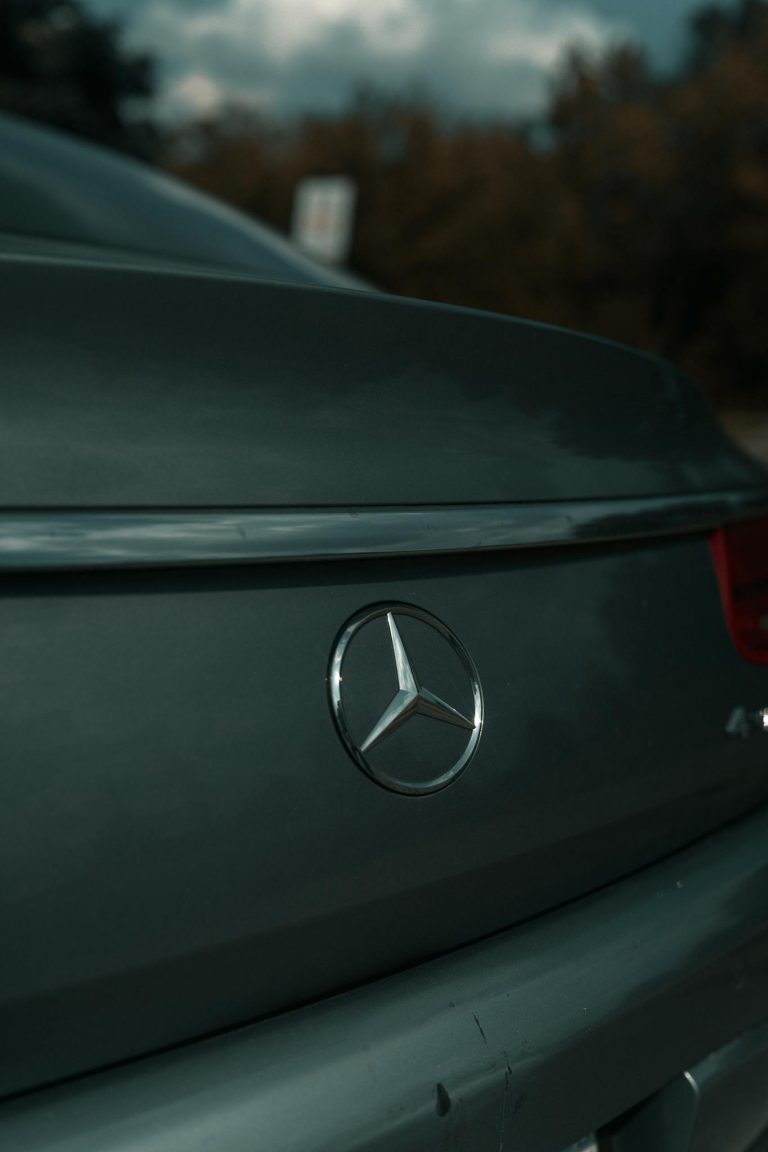 Close-up of Mercedes-Benz emblem on a luxury car's rear, surface details highlighted in natural light.